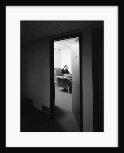 Executive businessman at desk seen working late through door ajar by Anonymous