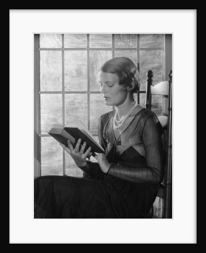Elegant woman wearing pearls sitting by window reading book by Anonymous