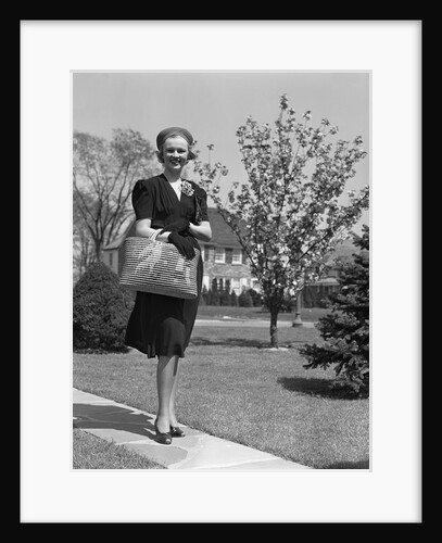 Stylishly dressed woman walking down suburban street carrying straw bag by Anonymous