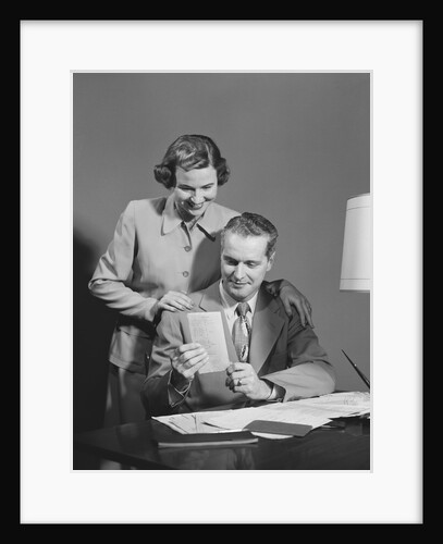 Couple man seated at desk holding paper woman standing looking over his shoulder smiling happy by Anonymous