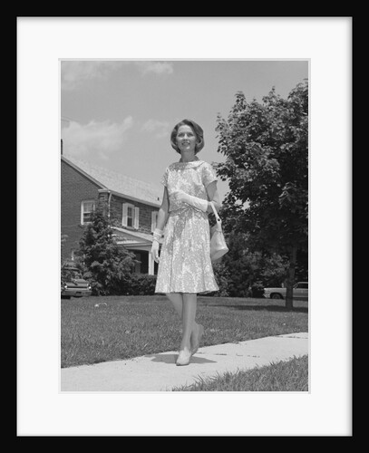 Woman wearing print dress holding handbag walking down suburban street by Anonymous