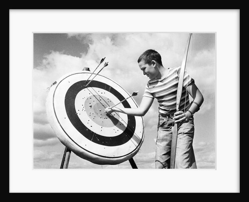 1960s boy jeans striped t-shirt holding bow & pulling arrow out of target bull's-eye by Anonymous
