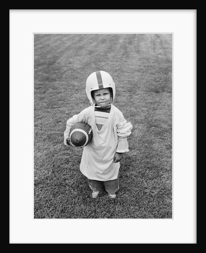 1950s boy standing in grass wearing oversized shirt & helmet holding football by Anonymous