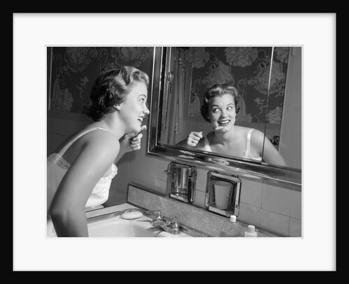 1950s smiling woman looking into bathroom mirror brushing teeth by Anonymous