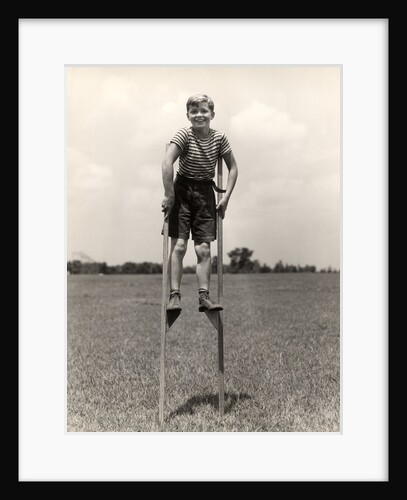 1930s 1940s smiling happy boy wearing striped shirt & short pants walking on pair of stilts looking at camera by Anonymous