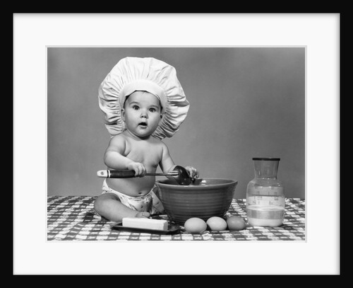 1960s baby seated on checkered tablecloth wearing chef's hat mixing eggs milk & flour in large bowl looking at camera by Anonymous