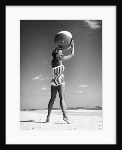 1960s woman in stripes swim suit bathing holding beach ball standing on tip toes by Anonymous