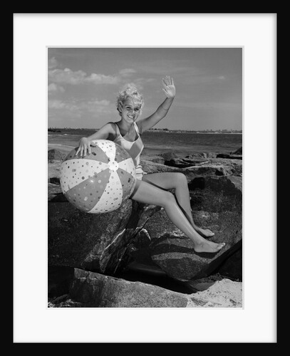 1960s smiling blond teenage girl sitting on rocky shore holding a beach ball waving looking at camera by Anonymous