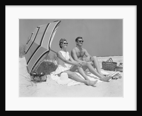 1960s couple in sunglasses sitting on beach blanket with legs extended with umbrella by Anonymous