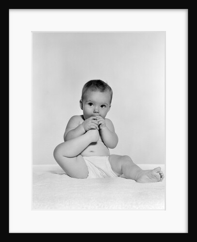 1950s 1960s baby seated on blanket bringing foot to mouth by Anonymous