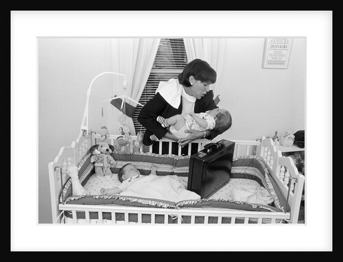 1980s working mother standing over crib holding one baby while other sleeps briefcase in crib indoor by Anonymous