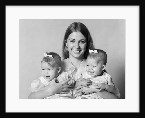 1970s portrait of smiling mother with arms around twin girls with bows in hair looking at camera by Anonymous