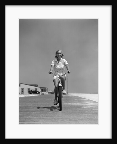 1940s summer time smiling blonde woman riding bike on seashore beach boardwalk directly looking at camera by Anonymous
