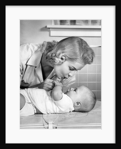 1950s mother woman bending over baby lying on layette holding hands talking by Anonymous