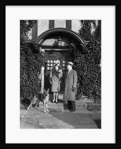 1920s couple wearing coat hat gloves on steps ivy covered building with german shepherd dog on a leash by Anonymous