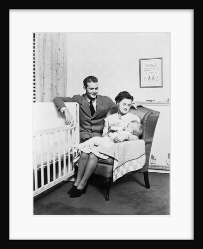 1930s 1940s mother sitting in chair in nursery baby 11 months on lap drinking from bottle father looking on leaning on crib by Anonymous