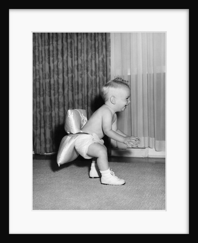 1950s laughing baby in diaper and shoes learning to walk with a pillow tied to his rear end by Anonymous