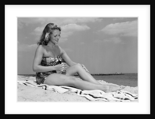 1950s woman in strapless one-piece bathing suit seated on beach towel putting on suntan lotion by Anonymous