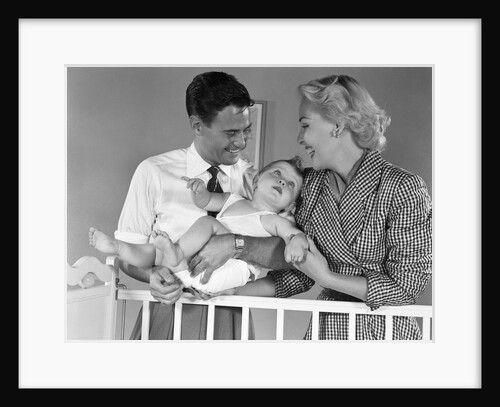 1950s smiling parents picking baby up out of crib by Anonymous