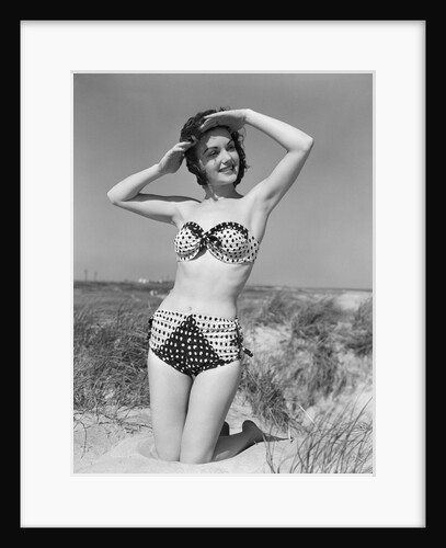 1950s smiling young woman kneeling in grassy sand wearing polka dot bikini shading eyes from sun by Anonymous