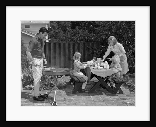 1960s man on patio grilling steak with 2 daughters seated at picnic table & wife standing serving food by Anonymous