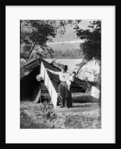 1920s mother holding baby on hip as she checks laundry diapers hanging on the line in front of tent by lake by Anonymous