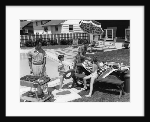 1960s family of 4 in backyard at poolside father barbecuing & mother & children making preparations at picnic table by Anonymous