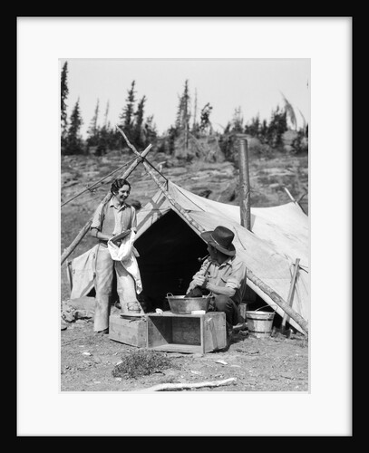 1930s smiling talking couple working by rustic western campsite tent man in cowboy hat smoking pipe washing skillet woman drying dishes by Anonymous