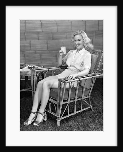 1940s blonde woman in bamboo chair holding up glass of milk smiling wearing white shorts looking at camera by Anonymous