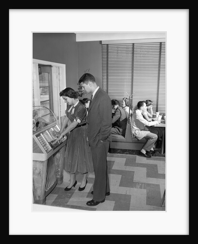 1950s teen couple playing juke box in malt shop with other teens in booths by Anonymous