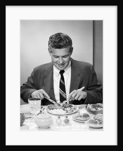 1950s smiling man in suit and tie sitting at table holding knife and fork eating dinner by Anonymous