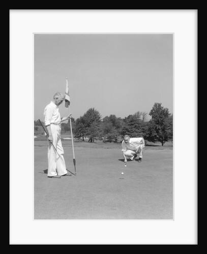 1930s 1940s elderly men on golf green one holding flag the other kneels lining up his putt by Anonymous