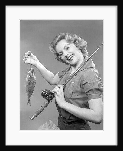 1950s smiling woman with a fishing rod over her shoulder holding up a fish looking at camera by Anonymous