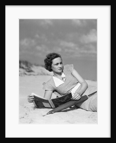 1930s woman leaning in wooden chair on beach wearing printed dress holding magazine looking thoughtful by Anonymous