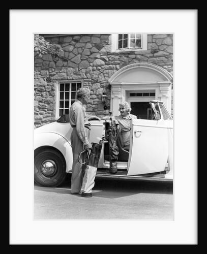 1940s senior retired couple loading golf clubs into white convertible car in front stone house by Anonymous