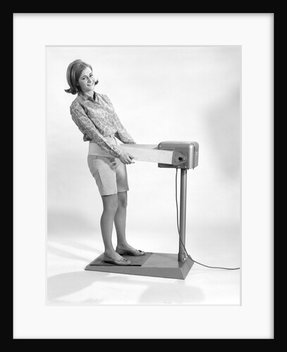 1960s smiling young woman standing on weight reducing vibrating exercise machine looking at camera by Anonymous