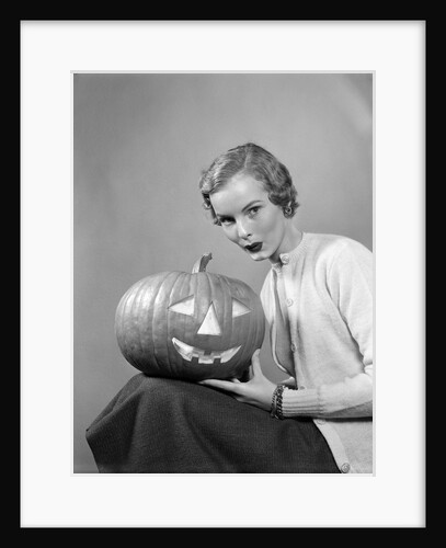 1950s woman wearing sweater holding carved pumpkin jack-o-lantern on her lap by Anonymous