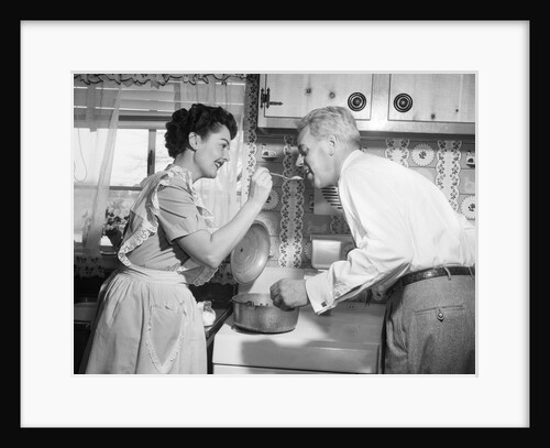 1950s housewife in kitchen having husband taste food on stove by Anonymous