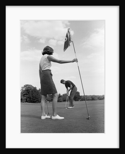 1970s couple playing golf man putting woman holding flag by Anonymous
