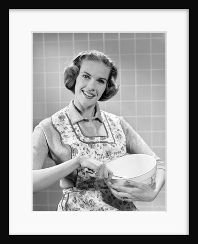 1950s smiling woman wearing apron in kitchen holding mixing bowl and spoon looking at camera by Anonymous