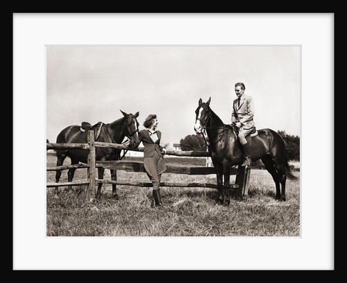 1930s 1940s couple in riding gear man riding horse woman standing by wooden fence by Anonymous