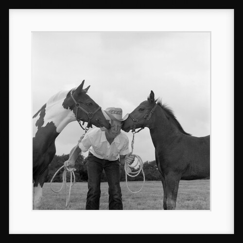 1960s boy wearing cowboy hat holding ropes reins harness halter of 2 horses one on either side of his face kissing him funny by Anonymous
