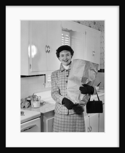 1950s smiling woman in kitchen holding grocery bag handbag wearing hat gloves looking at camera by Anonymous