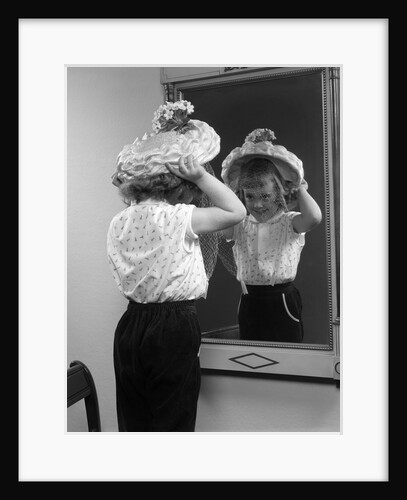 1950s little girl trying on hat looking into mirror reflection by Anonymous