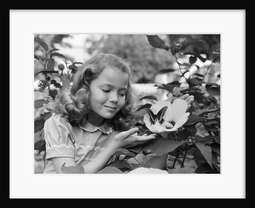 1950s pretty little girl smiling at hibiscus flower & butterfly in garden by Anonymous