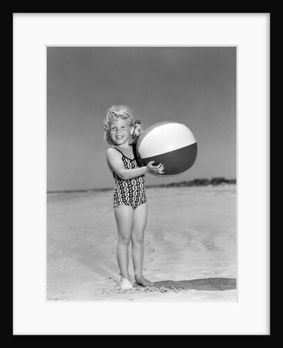 1950s smiling little girl standing on beach holding beach ball looking at camera by Anonymous