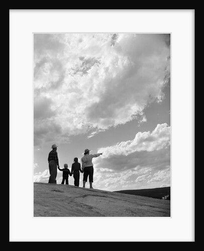 1950s 1960s family of four silhouetted on top rocky hill pointing at clouds in sky by Anonymous