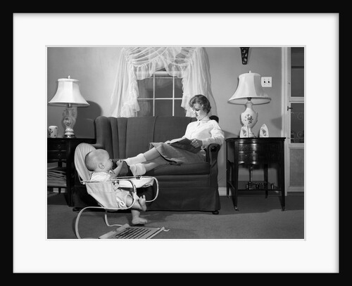 1950s teen babysitter seated on sofa reading schoolbook while baby in bouncy sling chair grabs her propped up foot by Anonymous
