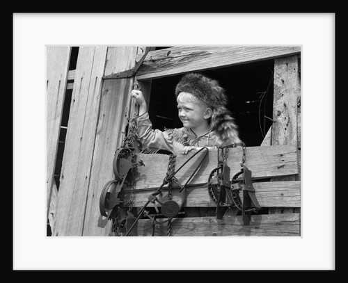 1950s boy dressed in coonskin hat & fringed vest like davy crockett outdoors in barn window by Anonymous