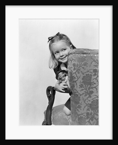 1940s little girl holding doll peeking around side of chair by Anonymous